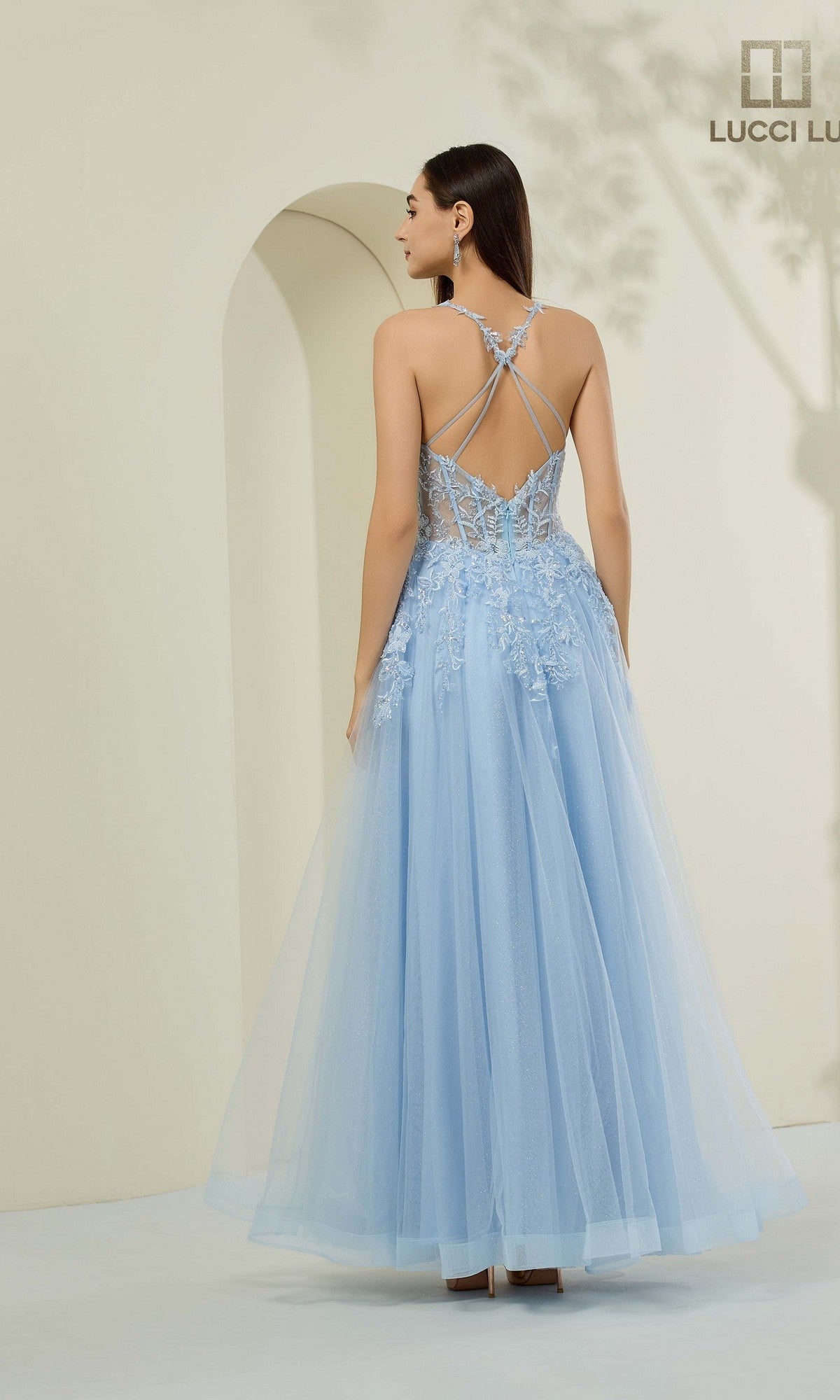 A woman stands facing away, wearing the Lucci Lu 1444 long prom dress with an a-line silhouette, corset bodice, light blue tulle skirt, lace details, and a strappy open back.